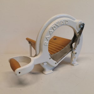 Puede incluir: Cortadora de pan vintage blanca con tabla de cortar y mango de madera. La palabra "BAADVAD" está grabada en el arco superior. La cuchilla es de acero inoxidable. Una herramienta de cocina clásica.