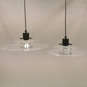 Könnte beinhalten: Zwei schwarze Pendelleuchten mit klaren, runden Acrylschirmchen. Die Leuchten sind an schwarzen Kabeln von der Decke abgehängt.