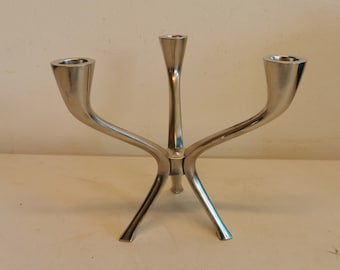 May include: A silver three-armed candlestick with a modern design. Each arm has a curved shape and ends in a cup for holding a candle.