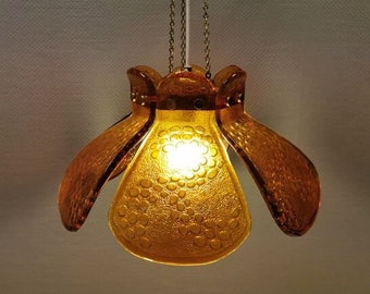May include: A gold-toned pendant light fixture with a flower-shaped shade made of amber glass. The shade has a textured surface with a pattern of small circles.