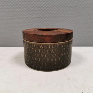 Può includere: Un piccolo contenitore rotondo in ceramica con un coperchio in legno marrone scuro. Il contenitore è di colore verde oliva tenue e presenta la parola "AKTOBAK" ripetutamente impressa all'esterno. Il coperchio ha un'apertura circolare al centro.