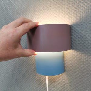 Puede incluir: Una l&aacute;mpara de pared moderna con una pantalla cil&iacute;ndrica. La parte superior es de color malva apagado, que pasa a azul claro en la parte inferior. La l&aacute;mpara emite una luz c&aacute;lida. Una mano sostiene la parte superior.
