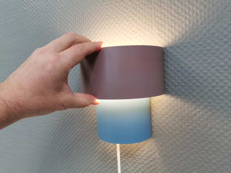 Puede incluir: Una l&aacute;mpara de pared moderna con una pantalla cil&iacute;ndrica. La parte superior es de color malva apagado, que pasa a azul claro en la parte inferior. La l&aacute;mpara emite una luz c&aacute;lida. Una mano sostiene la parte superior.