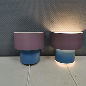 Puede incluir: Dos l&aacute;mparas modernas con un dise&ntilde;o cil&iacute;ndrico. Cada l&aacute;mpara tiene una parte superior de color lavanda y una base azul. Una l&aacute;mpara est&aacute; iluminada, emitiendo un brillo c&aacute;lido. Las l&aacute;mparas est&aacute;n conectadas a un cable de alimentaci&oacute;n blanco.