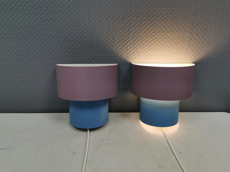 Puede incluir: Dos l&aacute;mparas modernas con un dise&ntilde;o cil&iacute;ndrico. Cada l&aacute;mpara tiene una parte superior de color lavanda y una base azul. Una l&aacute;mpara est&aacute; iluminada, emitiendo un brillo c&aacute;lido. Las l&aacute;mparas est&aacute;n conectadas a un cable de alimentaci&oacute;n blanco.