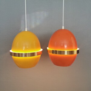 May include: Two hanging pendant lights with a gold metal band around the middle. The light on the left is yellow and the light on the right is orange.