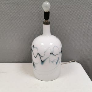 May include: White ceramic table lamp with a bulb. The lamp has a rounded base and a slender neck. The white surface is decorated with a flowing, abstract design in shades of blue and gray. The lamp is plugged into a white surface.