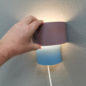 Puede incluir: Una l&aacute;mpara de pared moderna con una pantalla de dos tonos. La secci&oacute;n superior es de color p&uacute;rpura apagado, que pasa a un degradado azul en la parte inferior. Un cable blanco se extiende desde la base. La l&aacute;mpara est&aacute; montada en una pared texturizada.