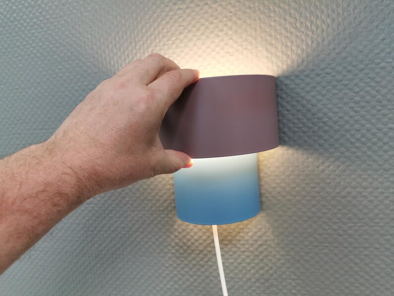 Puede incluir: Una l&aacute;mpara de pared moderna con una pantalla de dos tonos. La secci&oacute;n superior es de color p&uacute;rpura apagado, que pasa a un degradado azul en la parte inferior. Un cable blanco se extiende desde la base. La l&aacute;mpara est&aacute; montada en una pared texturizada.