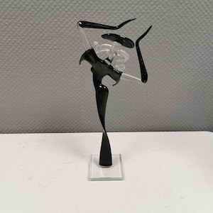 May include: A black and clear glass sculpture of a dancer in a graceful pose. The sculpture has a black body and limbs with clear glass accents. It stands on a clear square base, and the background is a light gray.