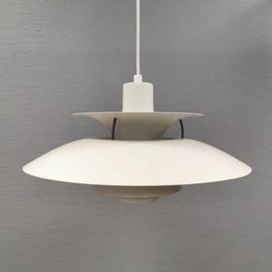 May include: A matte white pendant light fixture featuring a multi-tiered shade design with three stacked circular discs of varying sizes. The lamp is suspended by a single white cord attached to a central cylindrical housing with thin black supports.