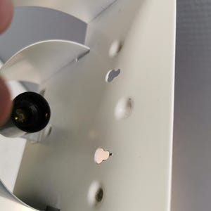 Puede incluir: Primer plano de una secci&oacute;n de un objeto met&aacute;lico blanco con varios agujeros peque&ntilde;os de forma irregular. Un objeto negro y redondeado con una punta dorada es visible en el lado izquierdo. El metal tiene algunas imperfecciones menores.