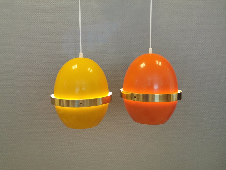 May include: Two hanging pendant lights shaped like eggs. One is yellow and the other is orange. Both have a gold metal band around the middle.