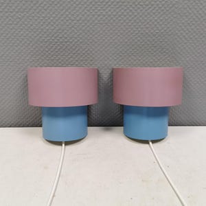 Puede incluir: Dos l&aacute;mparas de mesa modernas. Cada l&aacute;mpara tiene una base cil&iacute;ndrica azul y una pantalla rosa. Los cables blancos se extienden desde la base de cada l&aacute;mpara. Las l&aacute;mparas est&aacute;n sobre una superficie blanca contra una pared gris texturizada.