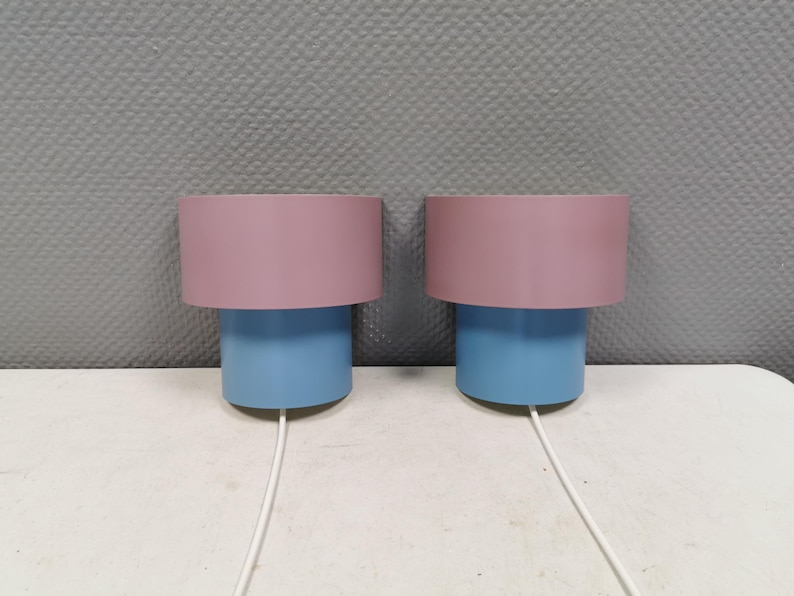 Puede incluir: Dos l&aacute;mparas de mesa modernas. Cada l&aacute;mpara tiene una base cil&iacute;ndrica azul y una pantalla rosa. Los cables blancos se extienden desde la base de cada l&aacute;mpara. Las l&aacute;mparas est&aacute;n sobre una superficie blanca contra una pared gris texturizada.