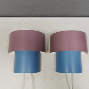Puede incluir: Dos apliques de pared a juego. Cada aplique tiene una base cil&iacute;ndrica azul y una pantalla semicircular malva. Las pantallas est&aacute;n montadas sobre un respaldo blanco. Los cables blancos se extienden desde la parte inferior de cada aplique.