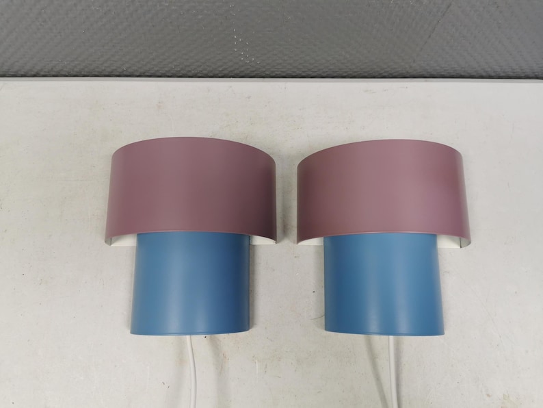 Puede incluir: Dos apliques de pared a juego. Cada aplique tiene una base cil&iacute;ndrica azul y una pantalla semicircular malva. Las pantallas est&aacute;n montadas sobre un respaldo blanco. Los cables blancos se extienden desde la parte inferior de cada aplique.