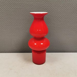 May include: A bright red glass vase featuring a stacked, multi-tiered silhouette with a cylindrical base, a rounded middle section, a bulbous upper body, and a flared rim. The interior of the rim is white, contrasting with the vibrant red exterior.