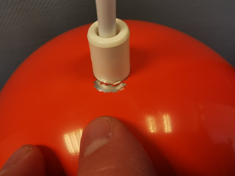 May include: Close-up of a red lampshade with a white cord and a silver metal ring around the top. The ring is slightly damaged.