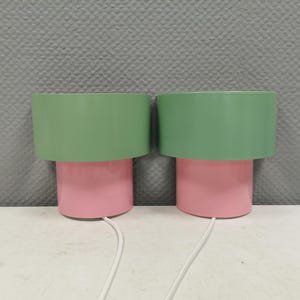 Puede incluir: Dos lámparas de mesa a juego. Cada lámpara tiene una base rosa cilíndrica y una pantalla verde cilíndrica ancha. Las lámparas están conectadas a un cable blanco. Las lámparas están sobre una superficie blanca con un fondo gris texturizado.