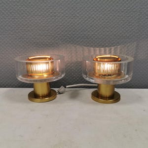 Pode incluir: Duas luminárias de mesa modernas. Cada luminária apresenta um abajur cilíndrico transparente e uma base dourada. As luminárias estão acesas, emitindo um brilho quente e convidativo. O design é minimalista e elegante, adequado para um interior contemporâneo.