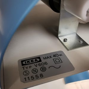 Puede incluir: Primer plano de una l&aacute;mpara de escritorio IKEA azul y blanca. La l&aacute;mpara tiene un soporte met&aacute;lico y una etiqueta con el logotipo de IKEA, el n&uacute;mero de modelo y las especificaciones de alimentaci&oacute;n. Dise&ntilde;o minimalista y moderno.