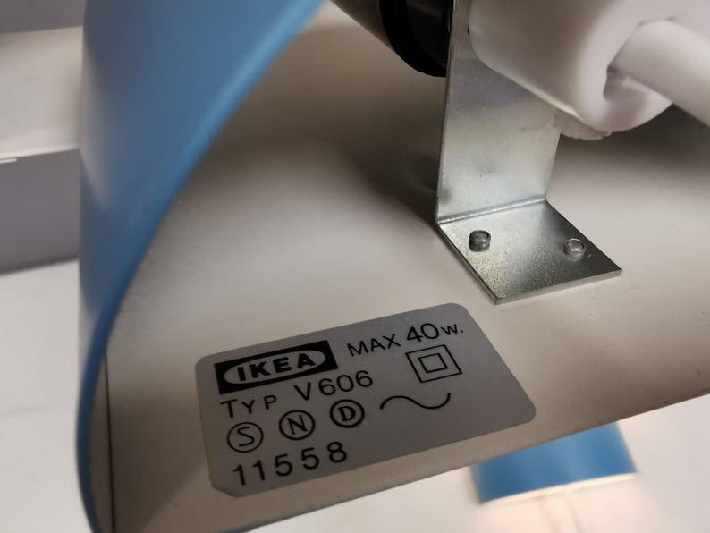 Puede incluir: Primer plano de una l&aacute;mpara de escritorio IKEA azul y blanca. La l&aacute;mpara tiene un soporte met&aacute;lico y una etiqueta con el logotipo de IKEA, el n&uacute;mero de modelo y las especificaciones de alimentaci&oacute;n. Dise&ntilde;o minimalista y moderno.