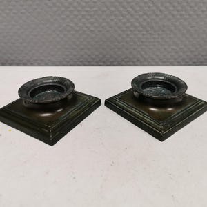 May include: Two small, square, black candle holders with a raised rim and a decorative design. Each holder has a small, round, silver-coloured candle cup.