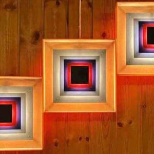 May include: Three wooden picture frames with a layered square design. Each frame has a black square in the center, surrounded by a red square, then a purple square, and finally a white square. The frames are mounted on a wood panel.