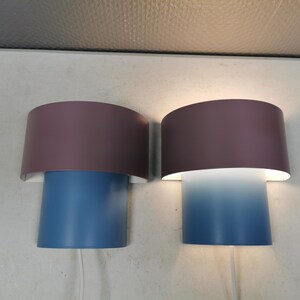 Puede incluir: Dos apliques de pared modernos. Cada aplique tiene una base cil&iacute;ndrica azul y una pantalla semicircular malva. Una pantalla est&aacute; encendida, proyectando un degradado de luz azul. Los apliques est&aacute;n montados en una pared blanca.