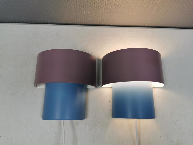 Puede incluir: Dos apliques de pared modernos. Cada aplique tiene una base cil&iacute;ndrica azul y una pantalla semicircular malva. Una pantalla est&aacute; encendida, proyectando un degradado de luz azul. Los apliques est&aacute;n montados en una pared blanca.