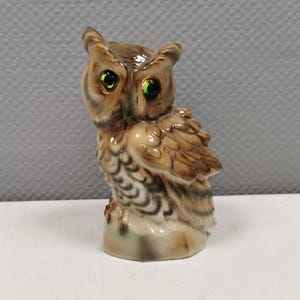 Può includere: Una piccola statuetta di gufo in ceramica con piume marroni e bianche. Il gufo ha grandi occhi verdi ed è appollaiato su una base.