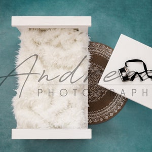 May include: White wooden crib with a white fluffy blanket inside. A brown and white patterned rug is on the floor next to the crib. A white square table with a black camera and strap is on the floor.