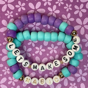 May include: Two beaded bracelets with the words "Let's Make Some Magic" spelled out in white beads with gold lettering. The beads are purple and turquoise.