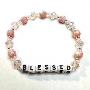 May include: A pink and white beaded bracelet with the word "BLESSED" spelled out in white letter beads.