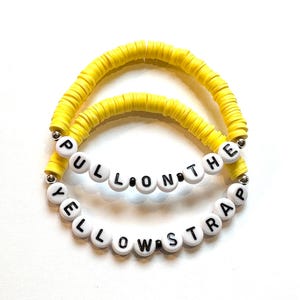 May include: Two yellow beaded bracelets. The top bracelet is made of yellow disc beads. The bottom bracelet has white letter beads that spell out "PULL ON THE YELLOW STRAP". The bracelets are on a white background.