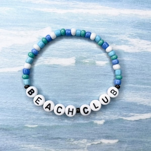 May include: A white beaded bracelet with blue and green beads spelling out "BEACH CLUB" in black letters.