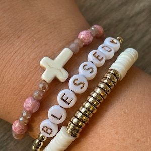 May include: A set of four bracelets featuring a white cross charm, a gold beaded bracelet, a white beaded bracelet with the word "BLESSED" spelled out in gold letters, and a pink and brown beaded bracelet.