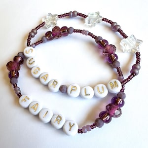 May include: Two beaded bracelets with the words "SUGAR PLUM" and "FAIRY" spelled out in gold letters on white beads. The bracelets feature purple and clear star-shaped beads.