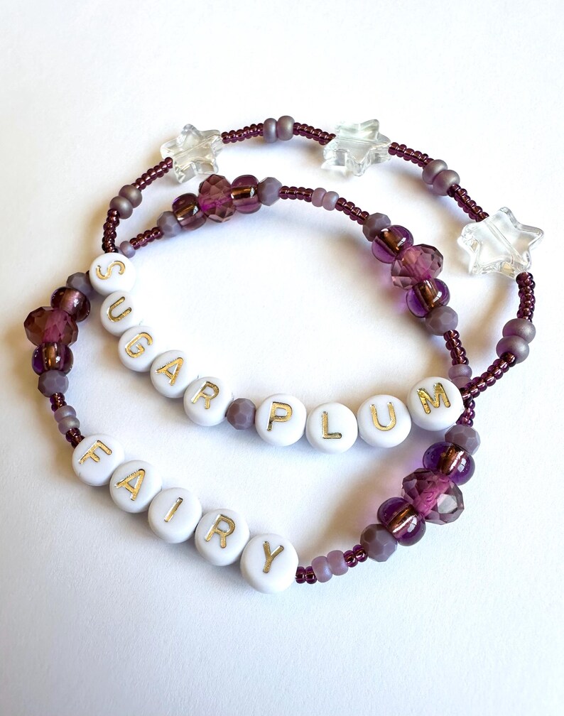 May include: Two beaded bracelets with the words "SUGAR PLUM" and "FAIRY" spelled out in gold letters on white beads. The bracelets feature purple and clear star-shaped beads.