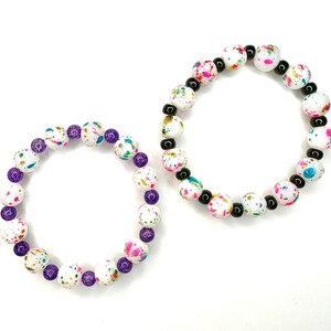 May include: Two beaded bracelets. One bracelet features white beads with colorful paint splatters and purple beads. The other bracelet has white beads with colorful paint splatters and black beads. Both bracelets are on a white background.