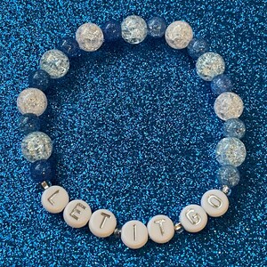 May include: A white beaded bracelet with blue and clear beads. The bracelet has the words "LET IT GO" spelled out on it.
