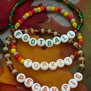 May include: Three beaded bracelets with the words "FOOTBALL", "TURKEY", and "PECAN PIE" spelled out in white letter beads. The bracelets are in fall colors of green, orange, yellow, and brown. The background features fall leaves.