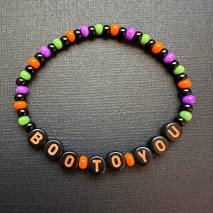 May include: A black beaded bracelet with orange, green, and purple beads. The bracelet spells out "BOO TO YOU" in orange letters.