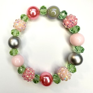 May include: A beaded bracelet featuring a variety of beads in shades of pink, silver, and green. The bracelet includes large, round beads, smaller faceted beads, and textured, sparkly beads. The beads are strung together with a clear elastic cord.