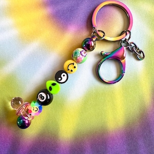 May include: A colorful keychain with a variety of beads and charms. The keychain features a rainbow-colored clasp, a large ring, and a series of beads with smiley faces, flowers, and other designs. The keychain also has a small pacifier charm.