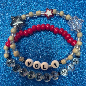 May include: Two beaded bracelets with the words "VOL 1 AWESOME MIX" on one and red and beige beads on the other. The bracelets are on a blue sparkly background.