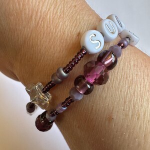 May include: Two beaded bracelets with purple and clear beads. One bracelet spells out the word "SOUL" with white letter beads. The other bracelet has a star-shaped charm and faceted purple beads. The bracelets are on a wrist.
