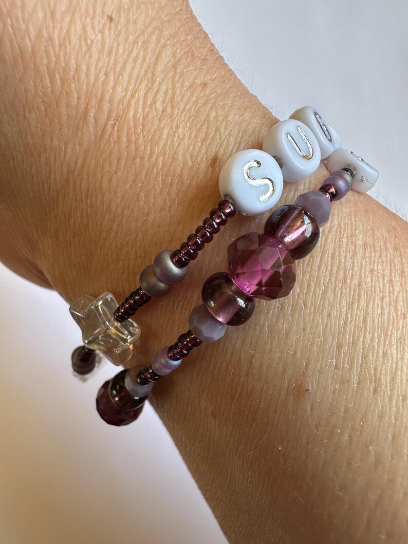 May include: Two beaded bracelets with purple and clear beads. One bracelet spells out the word "SOUL" with white letter beads. The other bracelet has a star-shaped charm and faceted purple beads. The bracelets are on a wrist.