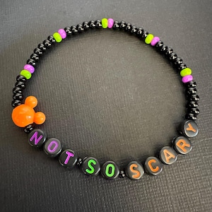 May include: A beaded bracelet featuring small black round beads interspersed with lime green and purple accents. An orange mouse-shaped bead is included, and black letter beads spell out the phrase NOT SO SCARY with purple, green, and orange lettering.